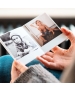Hands holding an open Polaroid-style photo book showing retro prints of children — one in black and white outdoors, and one playing indoors with a wooden spoon.